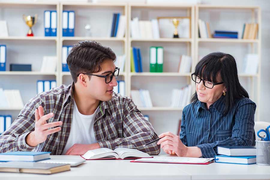Young man and older woman with glasses share a discussion over a textbook at a library desk.