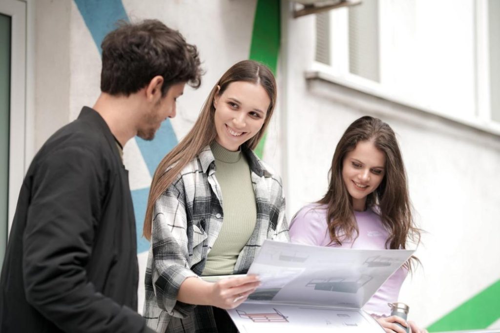 Three young adults examine a large blueprint together outdoors, smiling at its plans near a colorful wall mural.